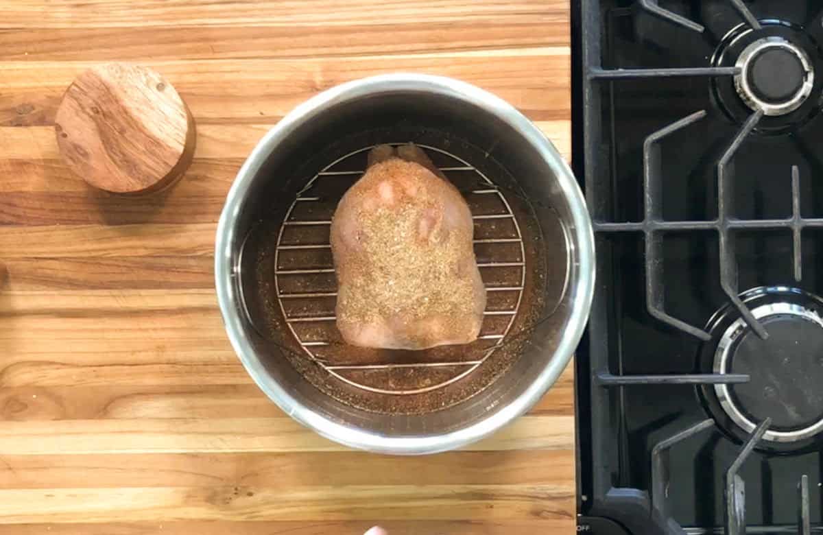 A seasoned piece of meat sits on a rack inside a metal pot on a wooden countertop, next to a stove with black grates. A round wooden container is also visible on the left.