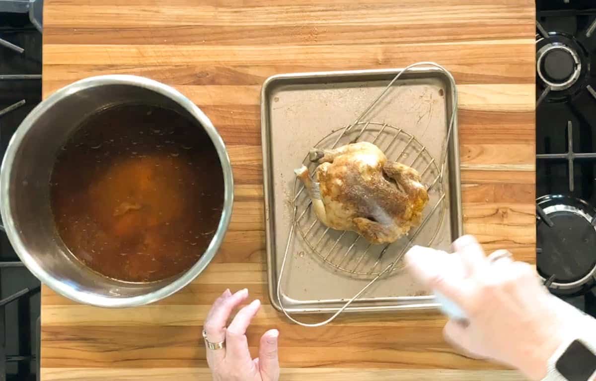 A person’s hand sprays liquid on a cooked whole chicken resting on a wire rack over a small baking sheet, with a pot of brown liquid nearby on a wooden countertop.