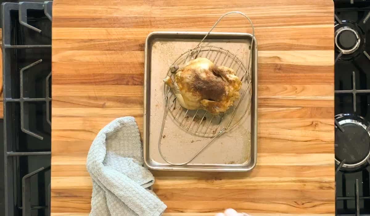 A whole roasted chicken sits on a wire rack placed over a baking sheet. Next to it is a folded gray kitchen towel on a wooden countertop, with parts of a stove visible on both sides.