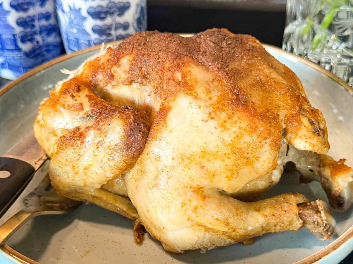 A whole roasted chicken with golden, seasoned skin sits on a plate. A knife and fork are partially visible to the side. Blue and white ceramic containers are in the background.
