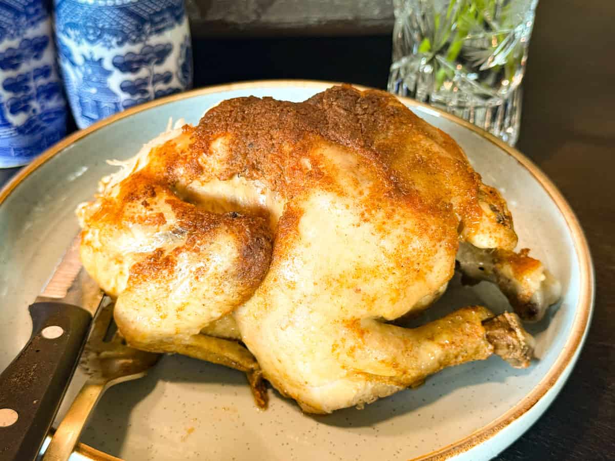 A whole cooked rotisserie chicken sits on a plate with a knife beside it. In the background, there are blue and white containers and a glass with green stems.