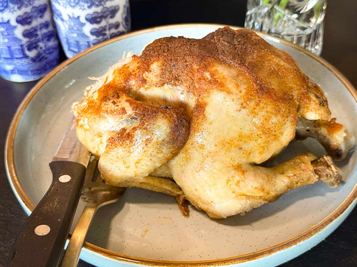 A whole roasted chicken with a browned, seasoned skin sits on a round plate. A carving fork and knife are placed beside it. Blue and white jars and a glass vase are partially visible in the background.