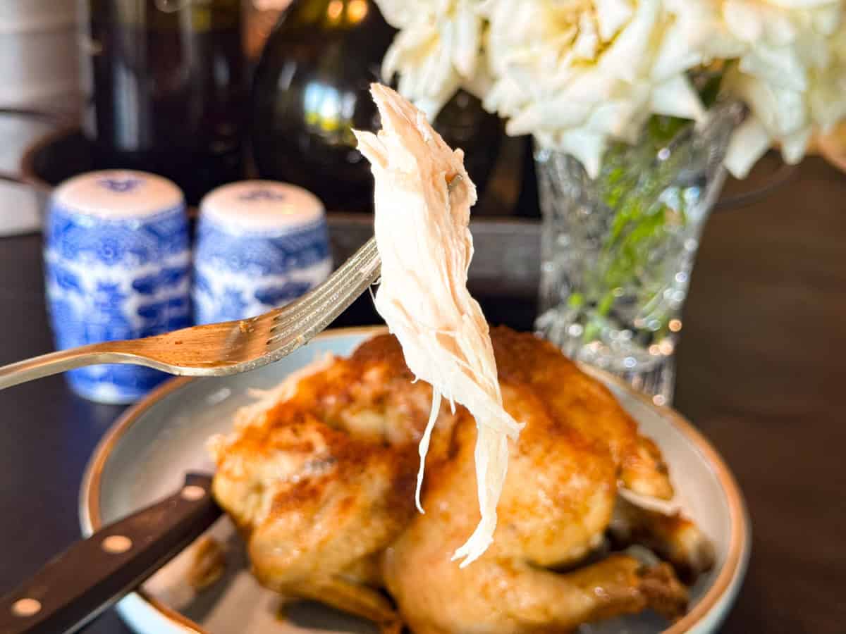 A close-up of a fork holding a piece of cooked chicken breast above a plate with a whole roasted chicken. In the background are blue and white salt and pepper shakers and a vase with white flowers.