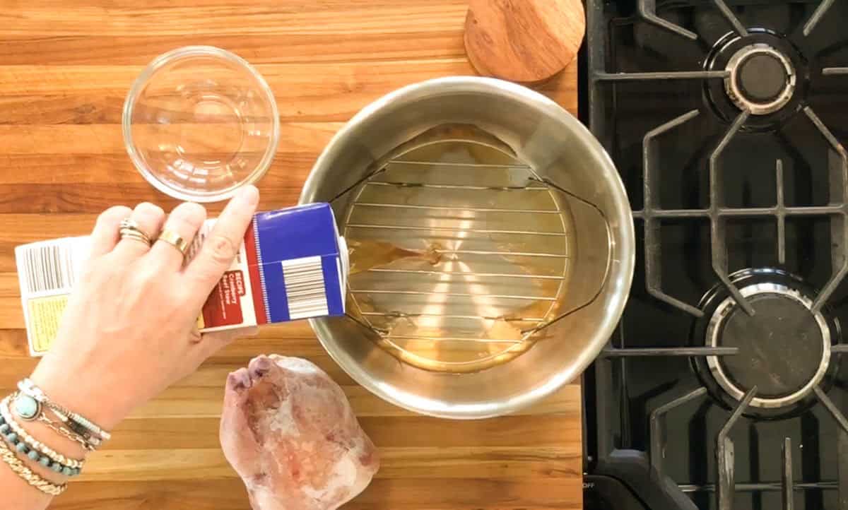 A hand pours liquid from a carton into a metal pot with a rack inside, placed on a wooden countertop next to a raw chicken and an empty glass bowl, beside a stovetop.