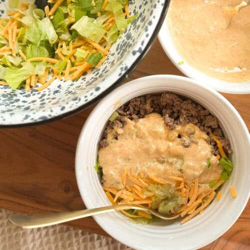 A bowl filled with ground beef, shredded cheese, lettuce, and a creamy sauce. Nearby are a bowl of lettuce with cheese and another bowl containing more sauce. A spoon rests in the main bowl.