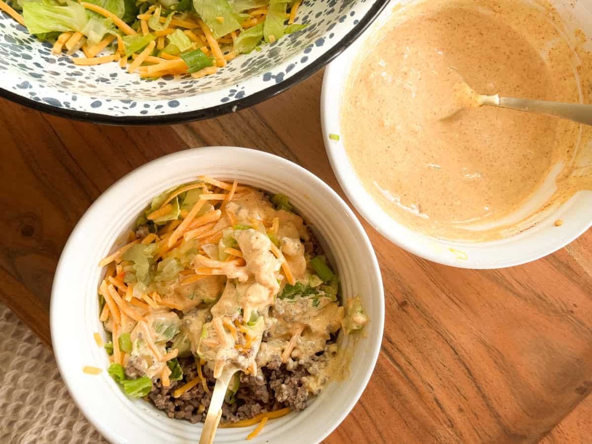 A bowl with ground beef, shredded cheese, lettuce, and sauce sits on a wooden table next to a large salad bowl and a smaller bowl of creamy dressing with a spoon.