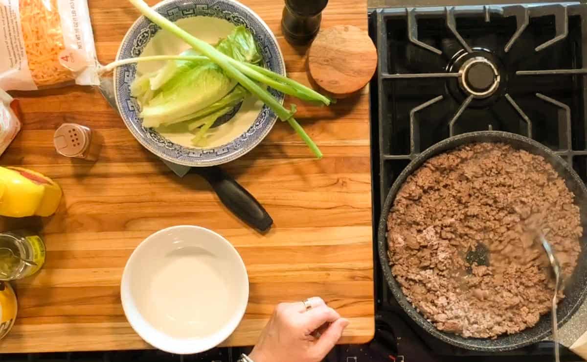 A person cooks ground beef in a skillet on a stove. A cutting board nearby holds lettuce, green onions, a knife, and a patterned bowl. Mustard, shredded cheese, and seasonings are also on the counter.
