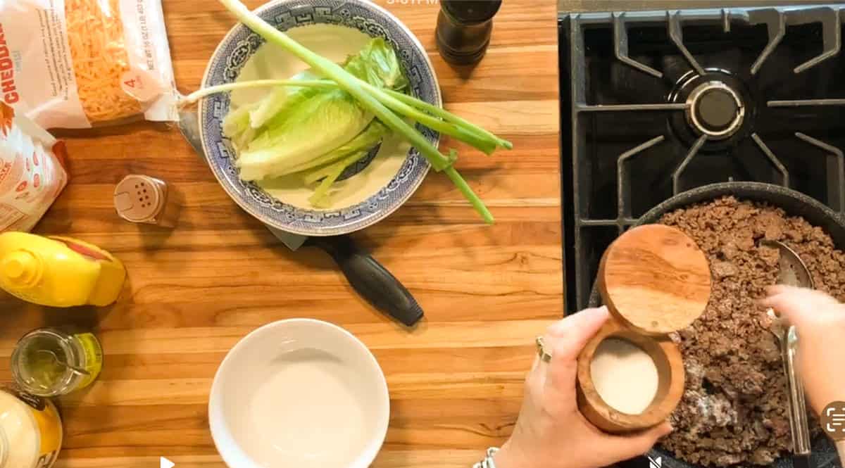 Overhead view of a person seasoning cooked ground beef in a skillet on a stove, with lettuce, green onions, grated cheese, and condiments arranged on a wooden countertop nearby.