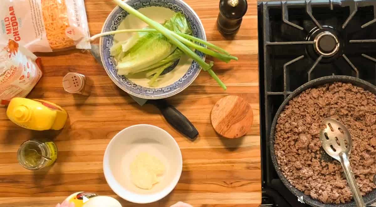A kitchen countertop with lettuce, green onions, cheese, mustard, mayonnaise, and seasonings next to a stove where ground beef is being cooked in a pan with a slotted spoon.