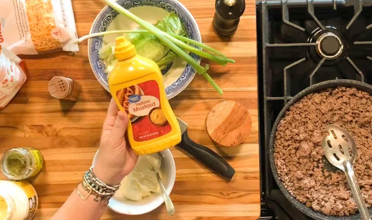 A hand holds a bottle of yellow mustard above a bowl of white sauce. Nearby are green onions, lettuce, shredded cheese, a spice shaker, mayonnaise, pickles, and a pan with cooked ground beef on a stove.
