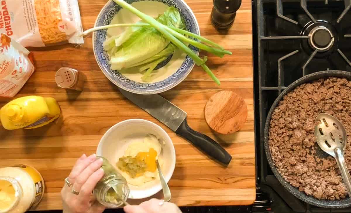 A person prepares sauce in a white bowl on a wooden counter with ground beef cooking in a skillet, lettuce and green onions in a bowl, shredded cheese, and condiments nearby.