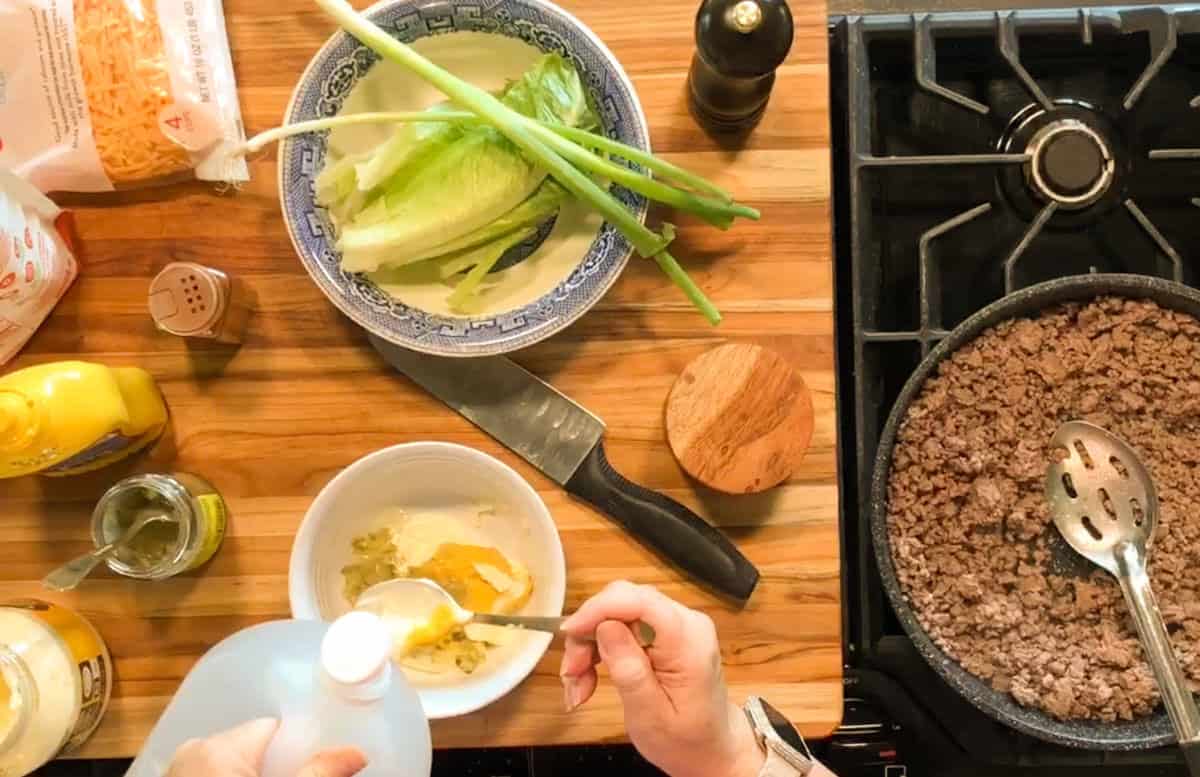Overhead view of a kitchen counter with chopped lettuce, green onions, and a knife on a plate, a bowl with condiments being mixed by hand, and a skillet of cooked ground beef next to a stove burner.