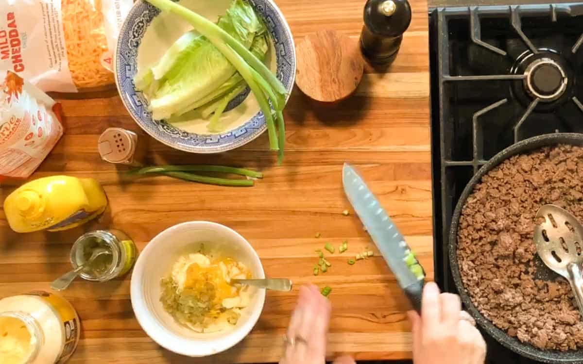 A person chops green onions on a wooden countertop next to a bowl of sauce, lettuce, cheddar cheese, green onions, condiments, and a skillet of cooked ground beef on a stove.