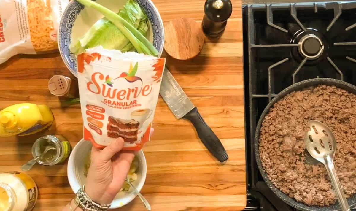 A hand holds a bag of Swerve granular sweetener above a wooden counter with green onions, lettuce, a knife, a bowl, condiments, and a pan of ground beef cooking on a stove.