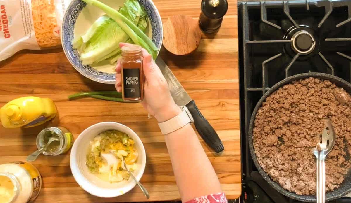 A person’s hand holds a jar of smoked paprika over a wooden countertop with lettuce, green onions, cheddar cheese, mustard, a knife, a bowl with mixed ingredients, and a skillet with cooked ground beef on a stove.