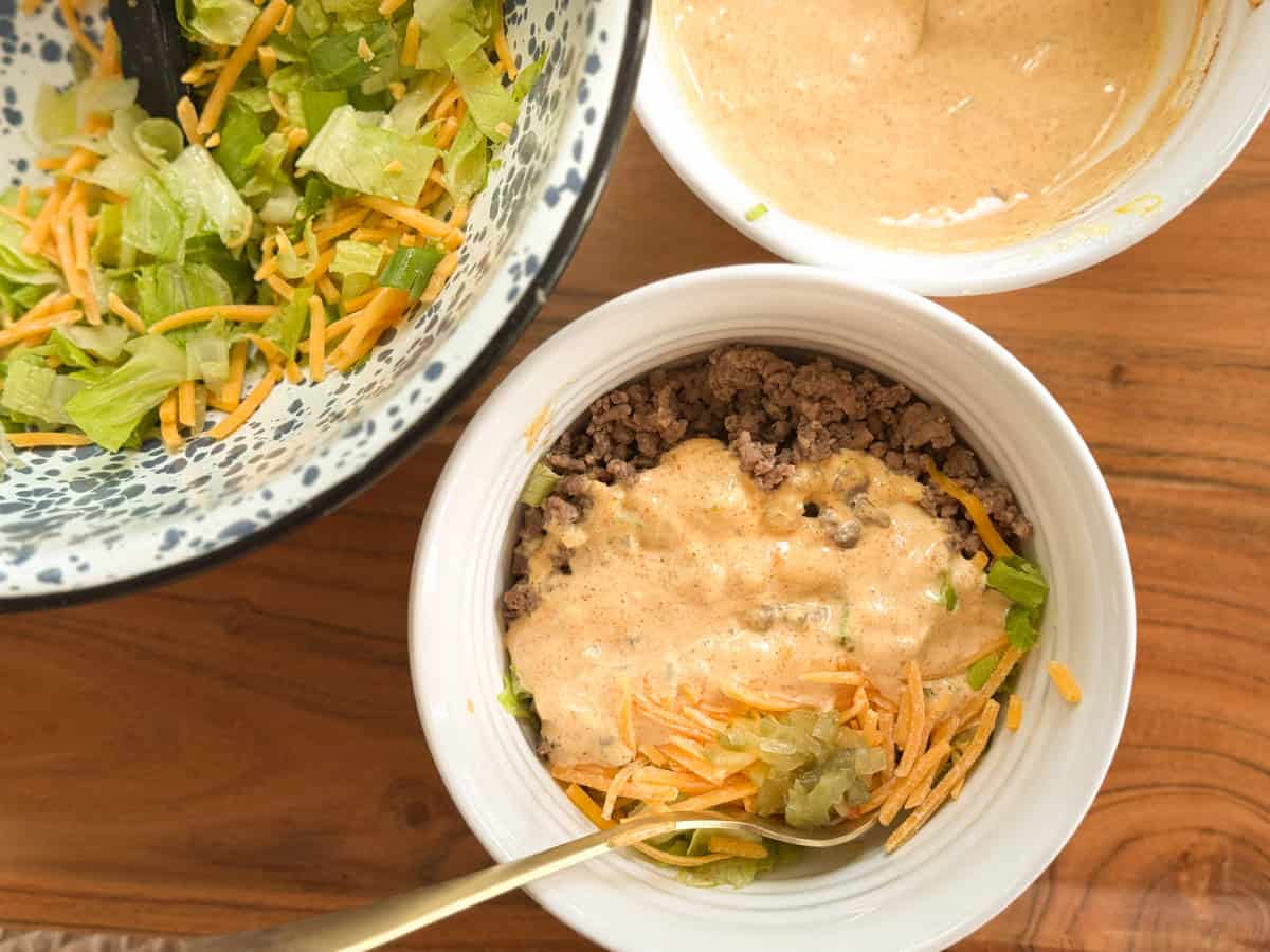 A bowl containing ground beef, shredded lettuce, shredded cheese, and a creamy sauce. Nearby are a bowl of lettuce with cheese and a bowl of creamy sauce, all set on a wooden surface.