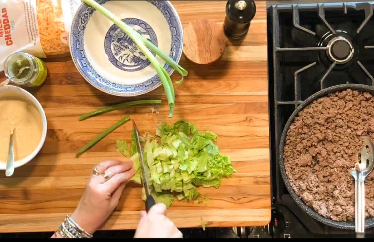 A person chops lettuce on a wooden cutting board next to a blue patterned plate, green onions, shredded cheese, a sauce, and a skillet of cooked ground beef on a stove.