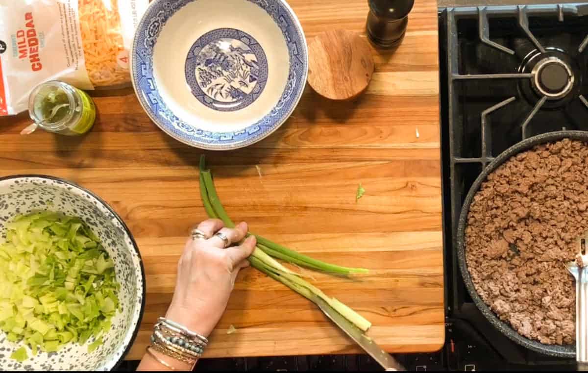 A hand holding green onions on a wooden cutting board, slicing them with a knife. There are bowls, a pepper grinder, shredded cheese, and cooked ground beef on the stove nearby.