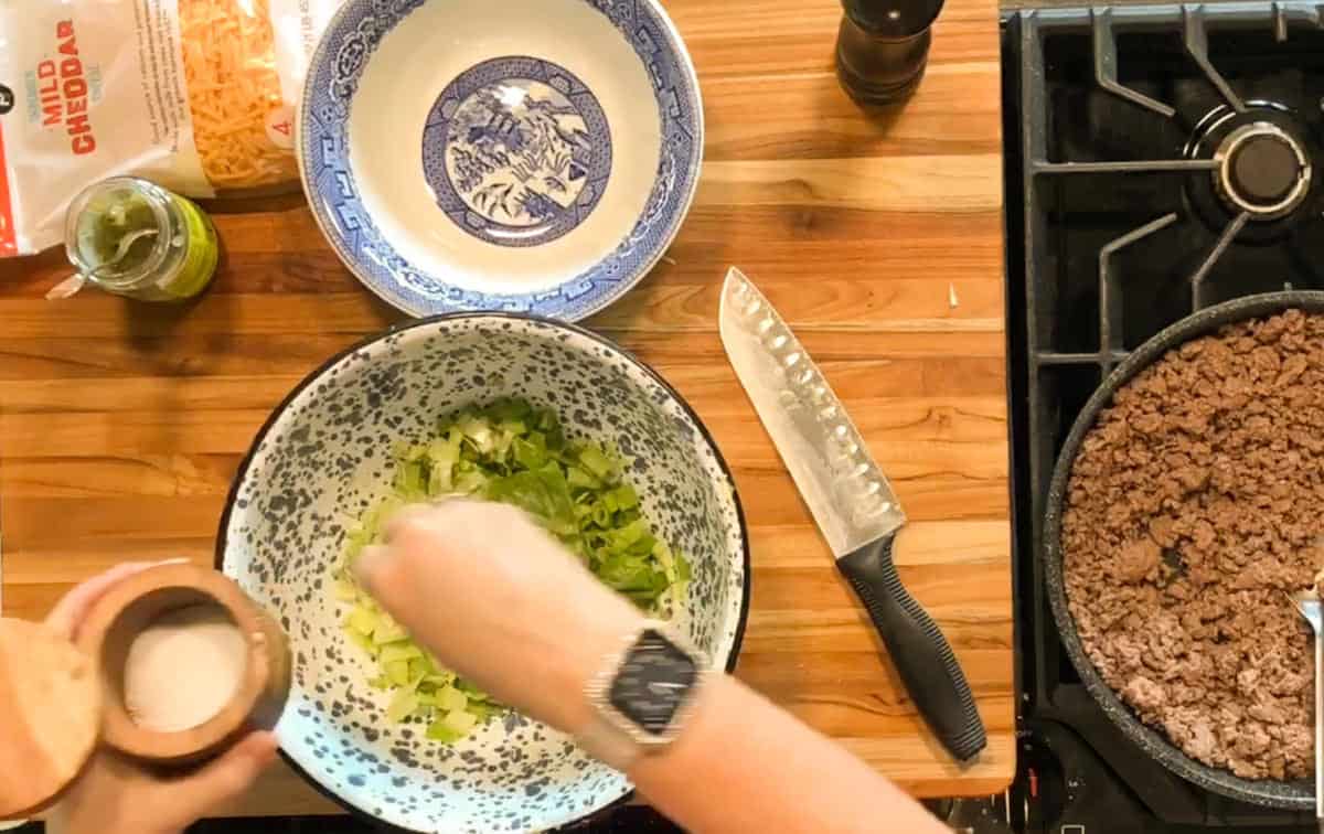 A person sprinkles salt into a large bowl of chopped green vegetables on a wooden countertop, with a knife, cooked ground beef in a pan, a bowl, shredded cheese, and a jar nearby.