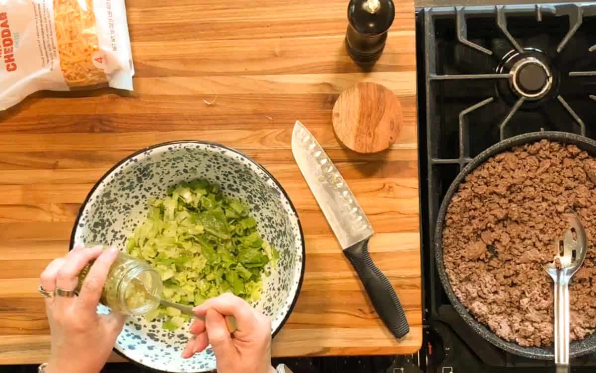 A person adds dressing to chopped lettuce in a large bowl beside a stove with cooked ground beef, a knife, pepper grinder, and a bag of shredded cheddar cheese on a wooden countertop.