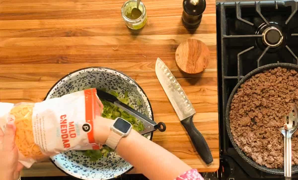 A person’s hand is pouring shredded mild cheddar cheese from a bag into a mixing bowl with green vegetables, next to a stove with cooked ground beef and utensils on a wooden countertop.