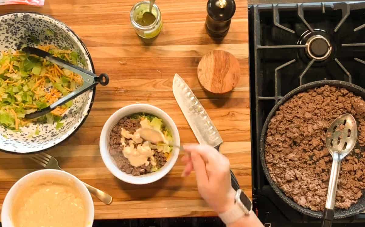 A person assembles a BIG MAC salad, adding sauce to a bowl of ground beef and lettuce. A pan with cooked beef, a salad bowl with cheese and lettuce, a knife, dressing, and seasonings are on a wooden countertop near a stove.
