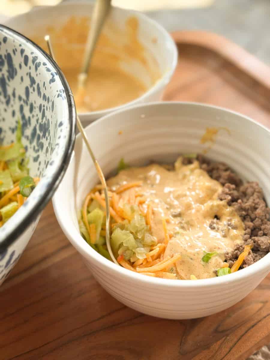 A white bowl on a wooden surface contains cooked ground beef, shredded lettuce, shredded cheese, pickles, and a creamy sauce. A second bowl with more sauce and a spoon is in the background.