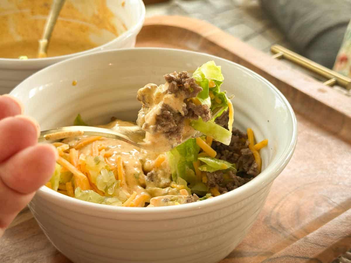 A hand holds a spoonful of salad with ground beef, lettuce, shredded cheese, and creamy dressing above a white bowl filled with the same ingredients on a wooden tray.