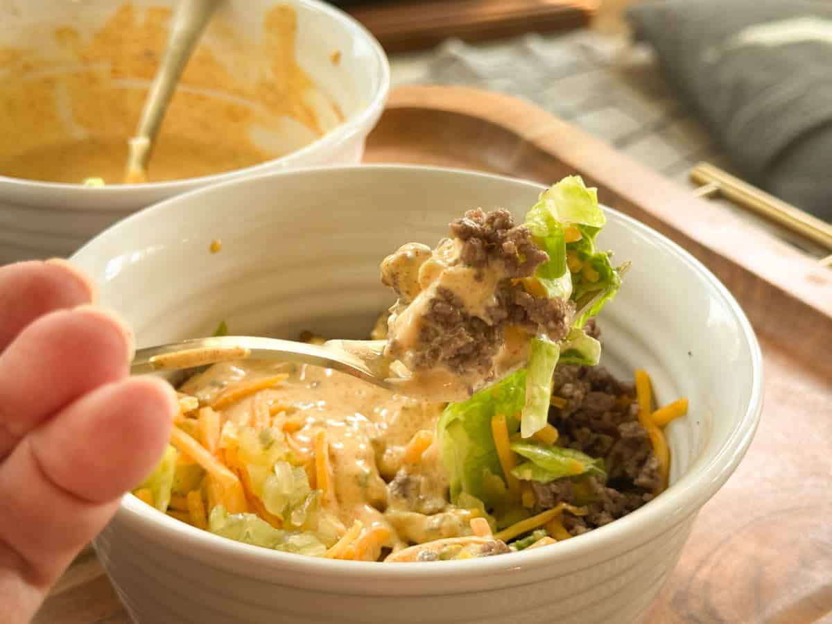 A hand holds a spoonful of a salad containing ground beef, shredded lettuce, cheese, and creamy dressing over a white bowl filled with the same ingredients. Another bowl and a tray are in the background.