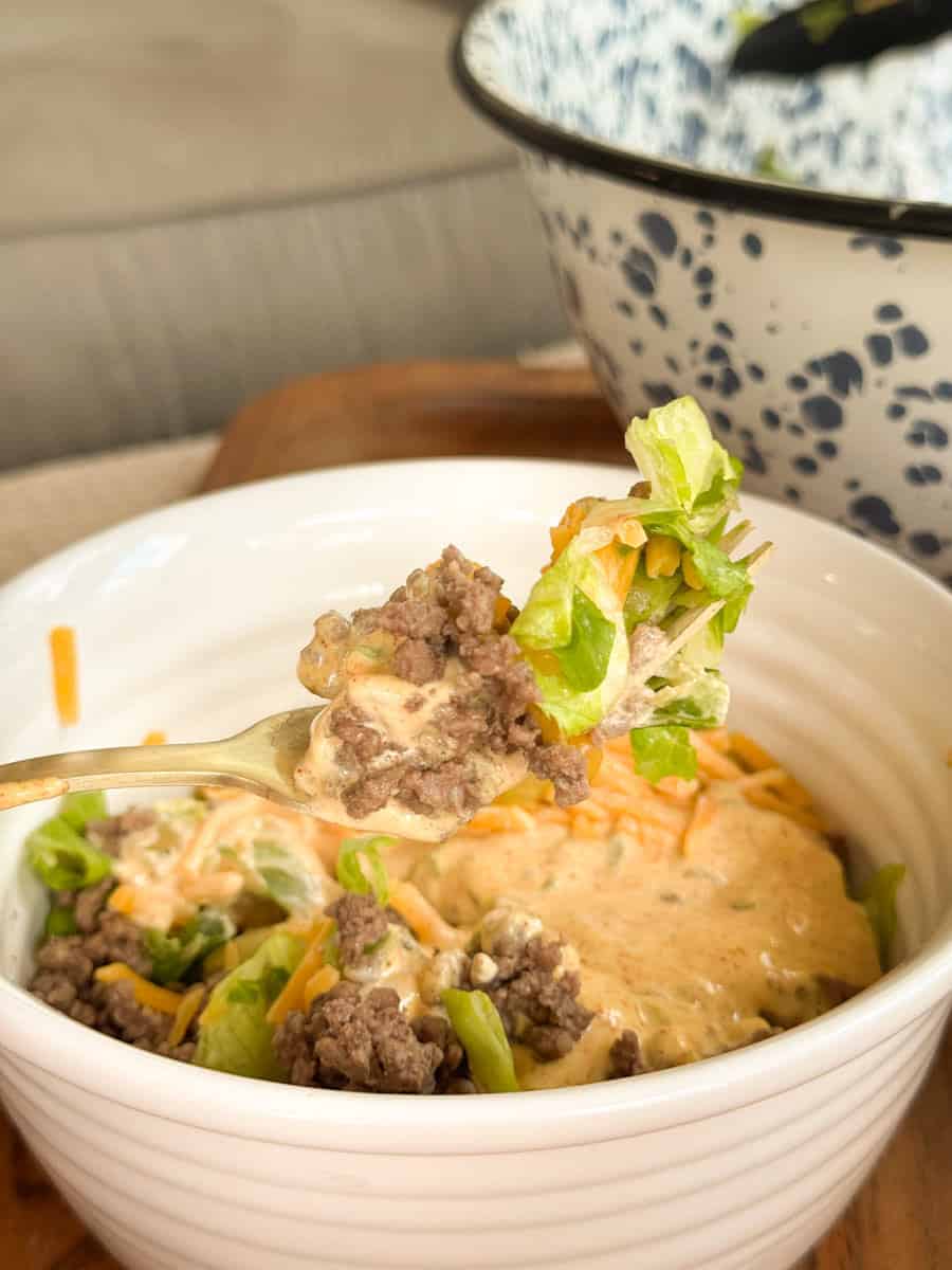 A fork holds a bite of ground beef, shredded cheese, lettuce, and creamy sauce over a white bowl filled with the same ingredients. A large, spotted bowl is in the background.