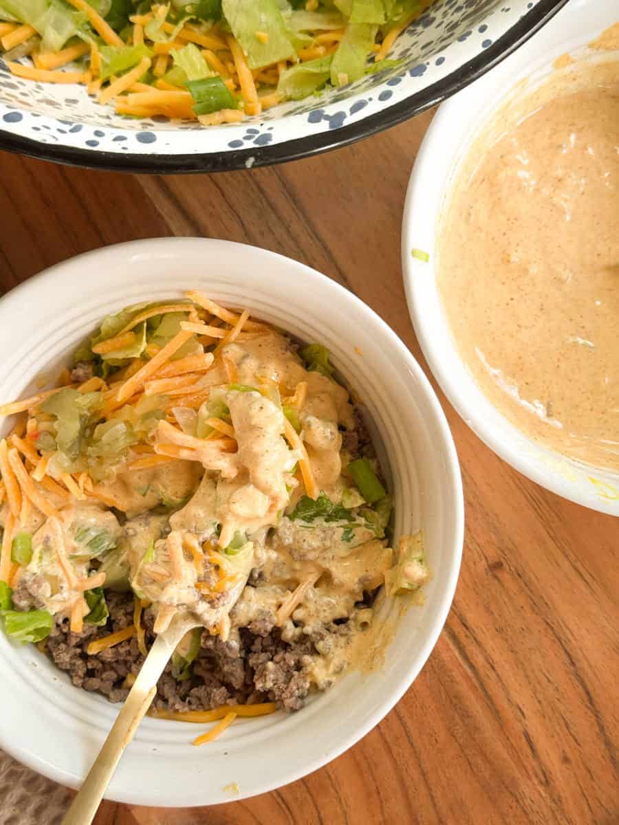 A white bowl filled with cooked ground beef, shredded lettuce, shredded cheese, and a creamy sauce sits on a wooden table. Nearby are two other bowls, one with sauce and one with more lettuce and cheese.