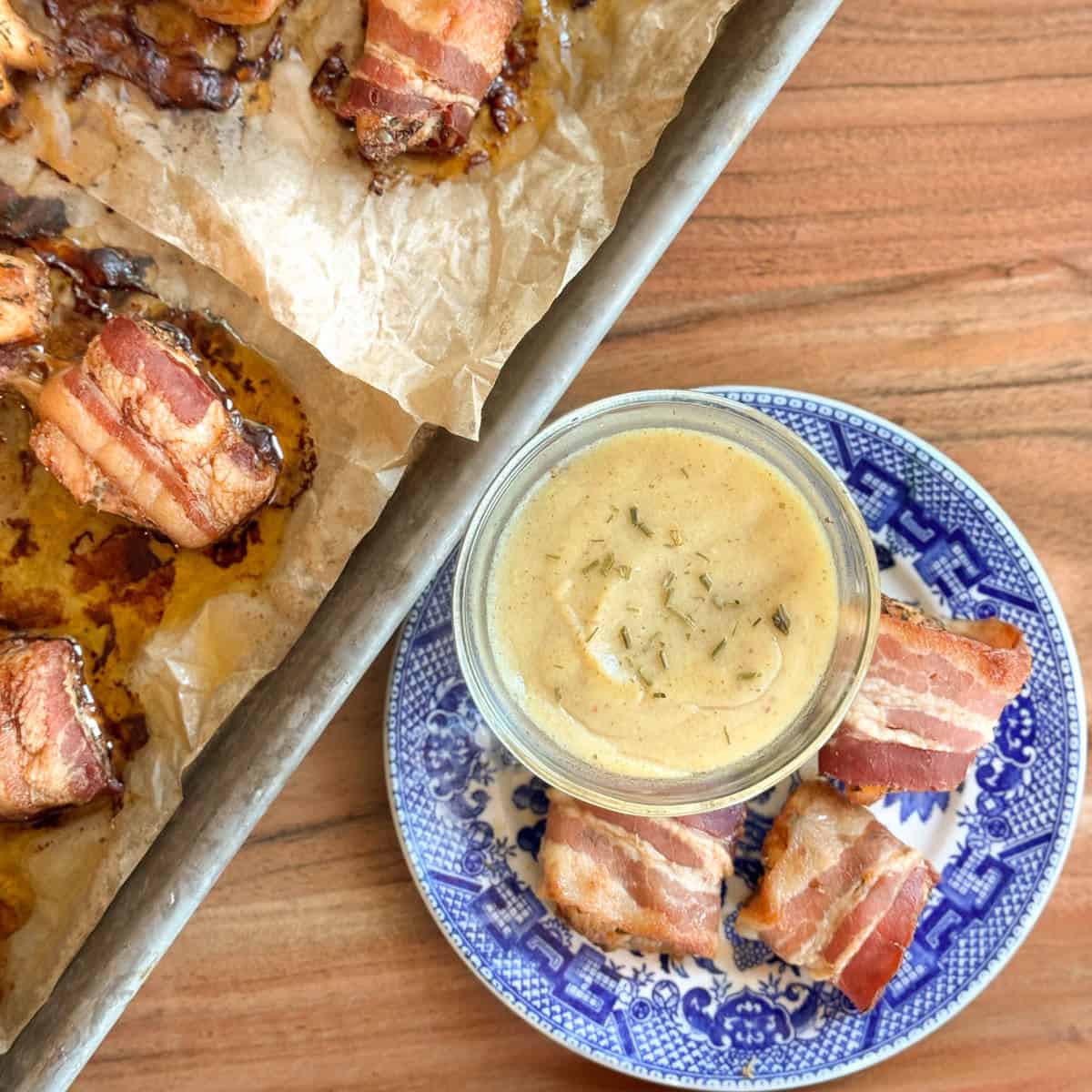 A tray with bacon-wrapped appetizers on parchment paper sits next to a blue and white plate holding three more bacon-wrapped pieces and a small glass jar of creamy dipping sauce topped with herbs.