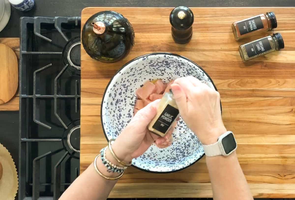 A person sprinkles garlic powder from a labeled jar onto raw chicken pieces in a speckled bowl on a wooden counter, with other spices, a pepper grinder, and an olive oil bottle nearby.