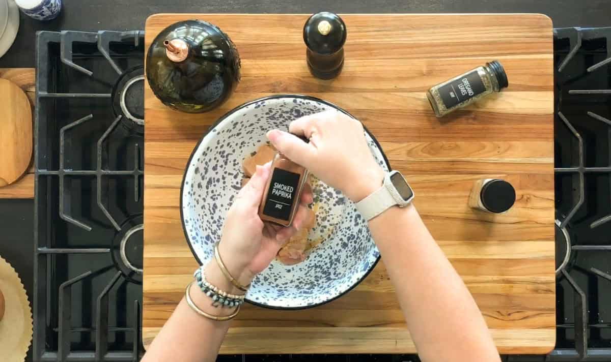 A person sprinkles smoked paprika from a jar onto raw chicken pieces in a large speckled bowl on a wooden cutting board. Other seasonings and a bottle of oil are nearby on the board.