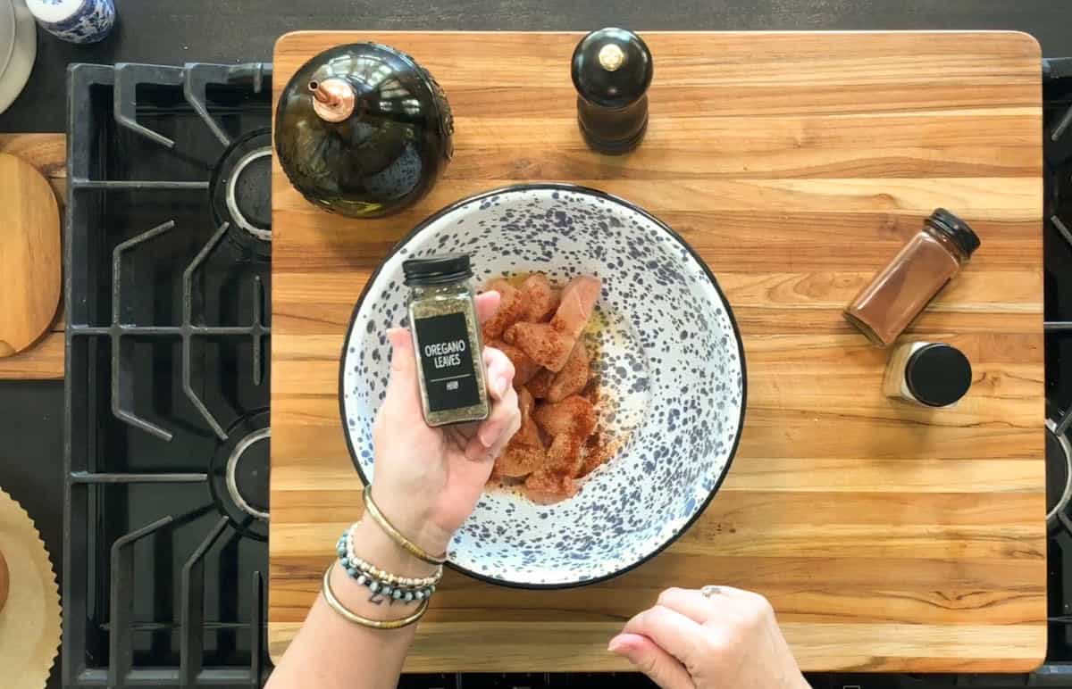 A person holds a jar labeled "oregano leaves" above a bowl of raw, seasoned chicken pieces on a wooden cutting board; spices and a pepper grinder sit nearby on a kitchen stove.