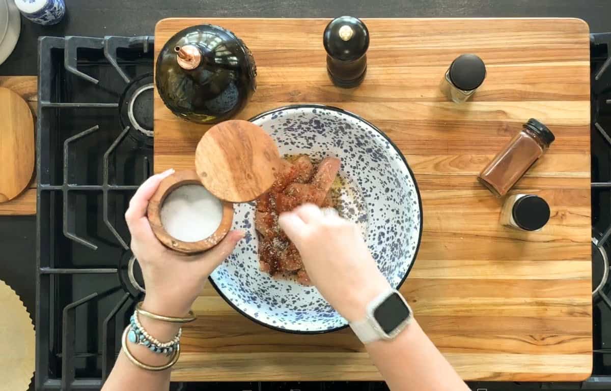 A person seasons raw chicken in a speckled bowl with salt, surrounded by spice containers, a pepper grinder, and a bottle on a wooden cutting board atop a stove.