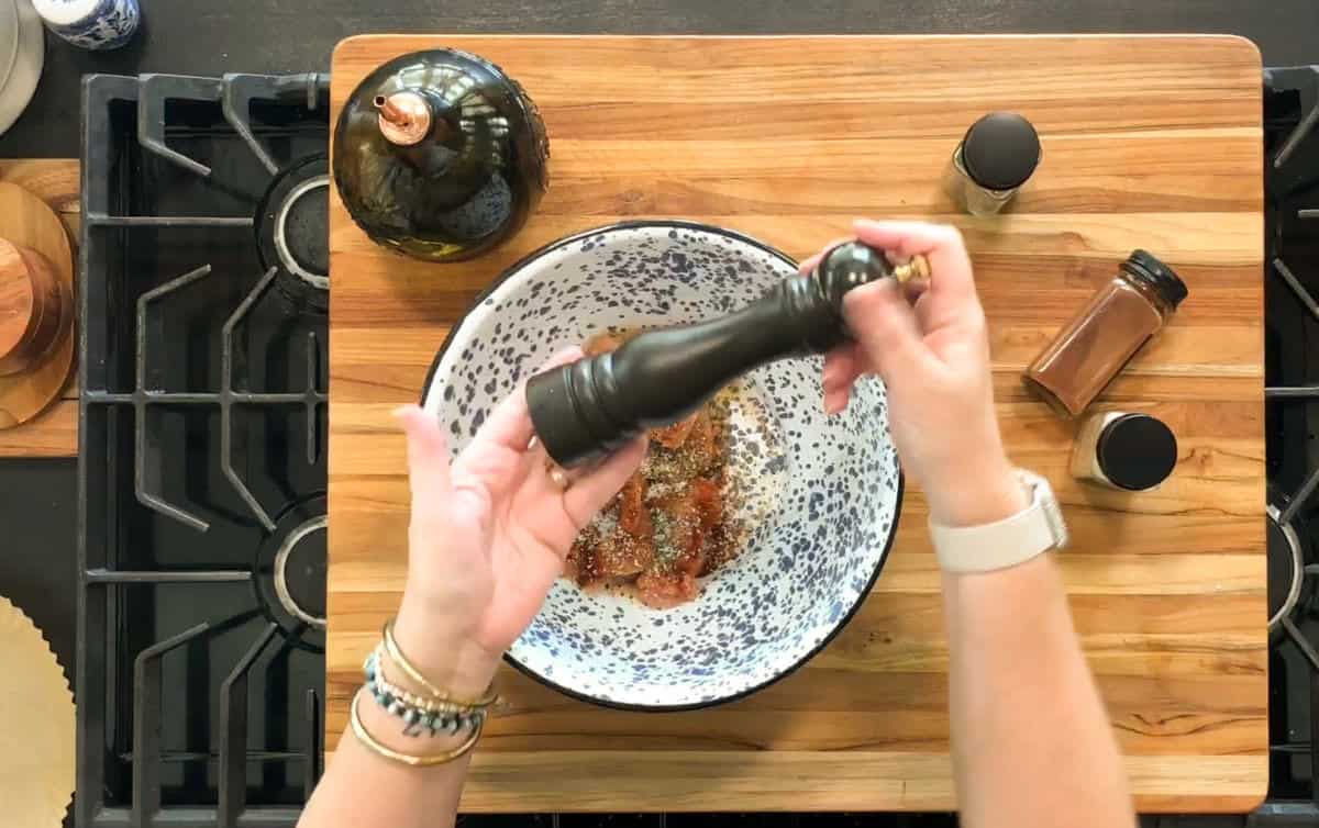 A person grinds black pepper over a bowl containing raw, seasoned meat. Spice jars and a ceramic container are on a wooden cutting board, which is placed above a stovetop.