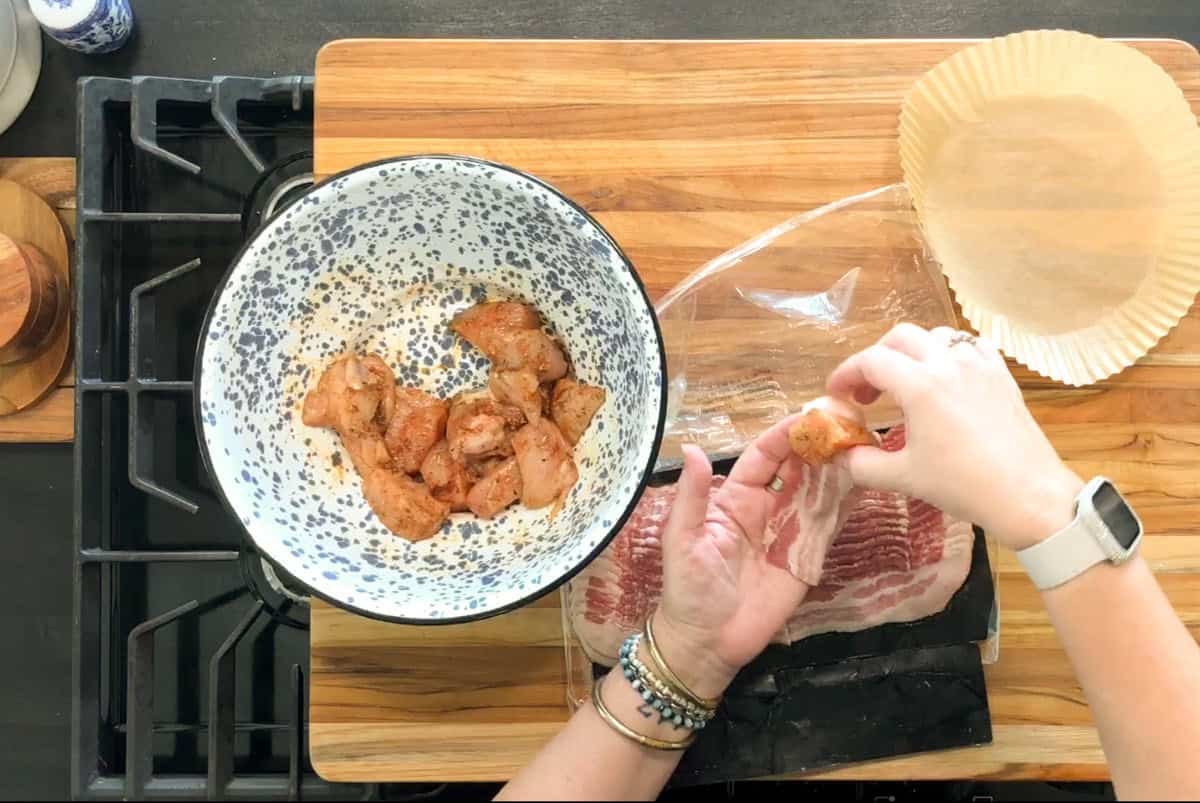 A person wraps a piece of seasoned chicken with bacon over a wooden cutting board. A bowl of chicken pieces, a tray of bacon, and a round parchment paper liner are also visible.