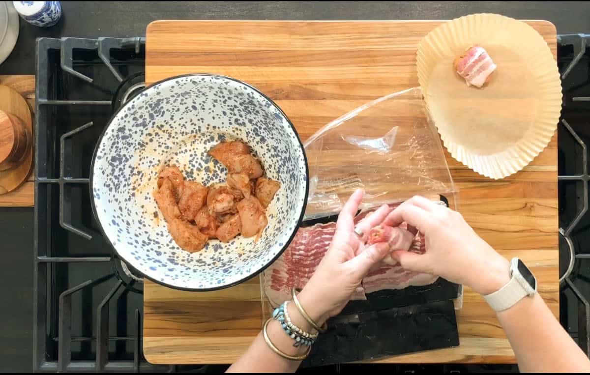 A person wraps a piece of seasoned chicken in bacon over a wooden cutting board. A bowl with more seasoned chicken is on the left, and bacon slices are on plastic wrap in the center.