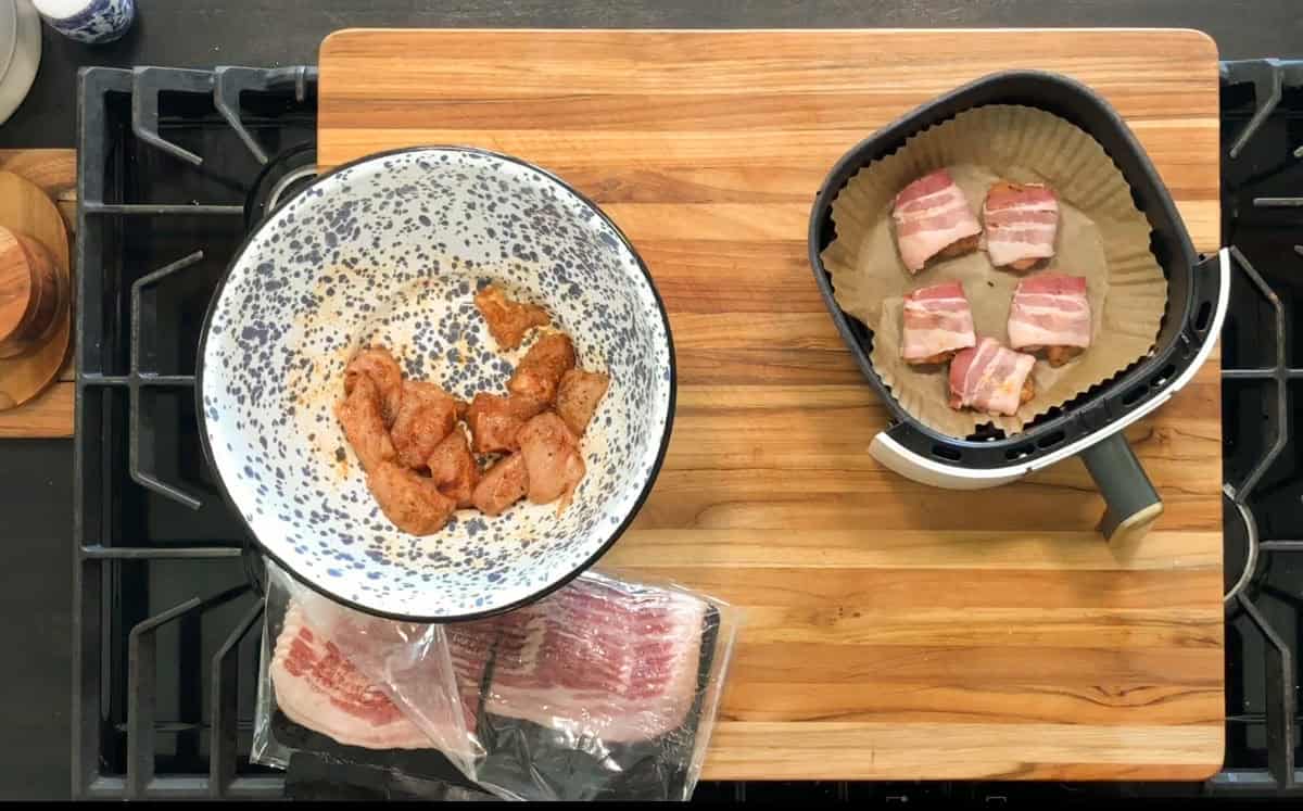 A bowl of seasoned chicken pieces sits on a wooden cutting board next to a package of bacon and an air fryer basket lined with parchment paper containing bacon-wrapped chicken pieces.