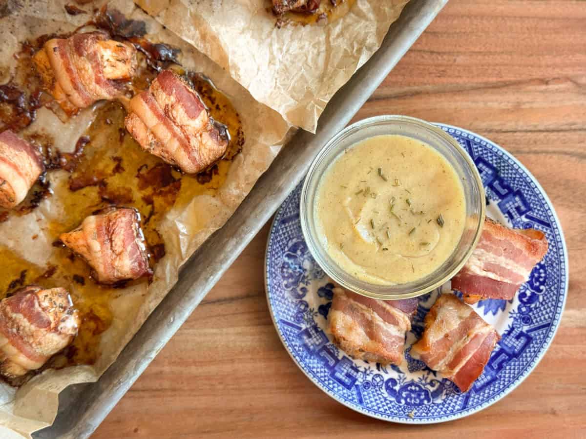 A tray with bacon-wrapped appetizers on parchment paper sits beside a blue and white plate holding a jar of dipping sauce and two bacon-wrapped pieces, all on a wooden surface.
