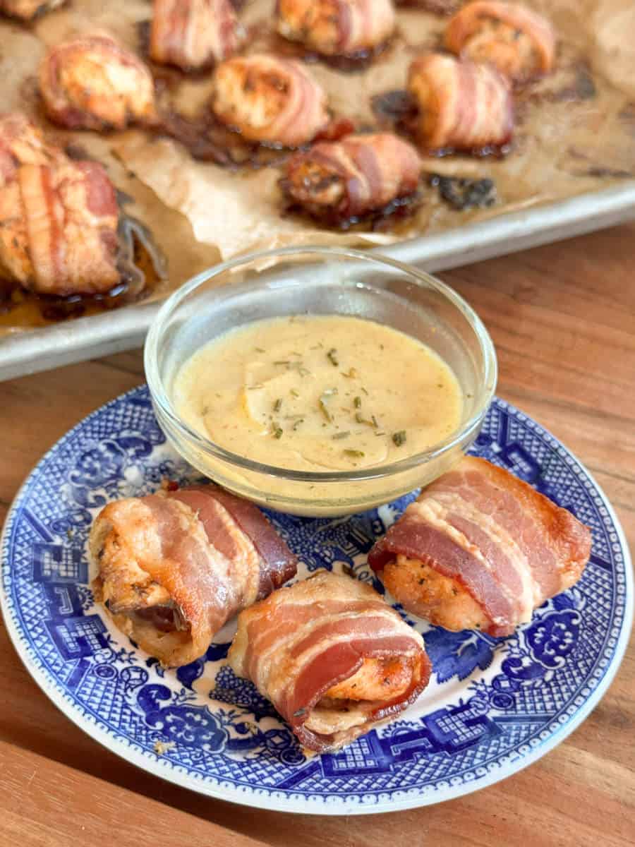 Three pieces of bacon-wrapped chicken are on a blue and white plate with a small bowl of creamy dipping sauce. More bacon-wrapped chicken pieces are visible on a parchment-lined baking sheet in the background.