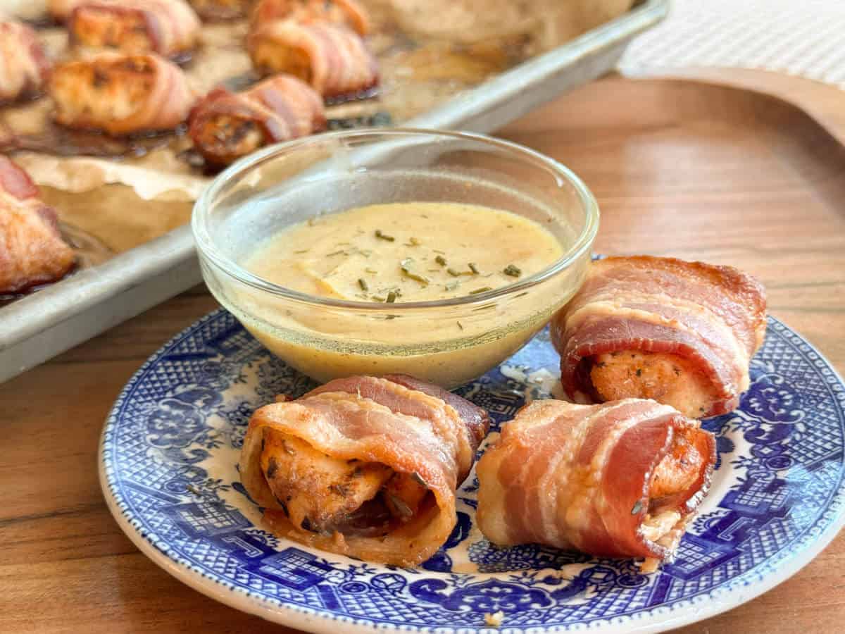 Three bacon-wrapped chicken bites are served on a blue and white patterned plate with a small bowl of creamy dipping sauce. A baking sheet with more bacon-wrapped bites is visible in the background.