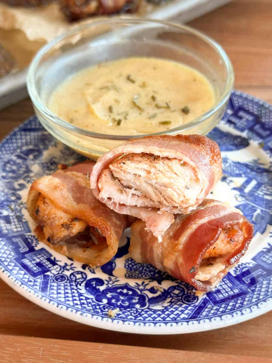 Three pieces of cooked bacon-wrapped chicken are arranged on a blue and white patterned plate, with a small glass bowl of creamy sauce and herbs placed behind them.