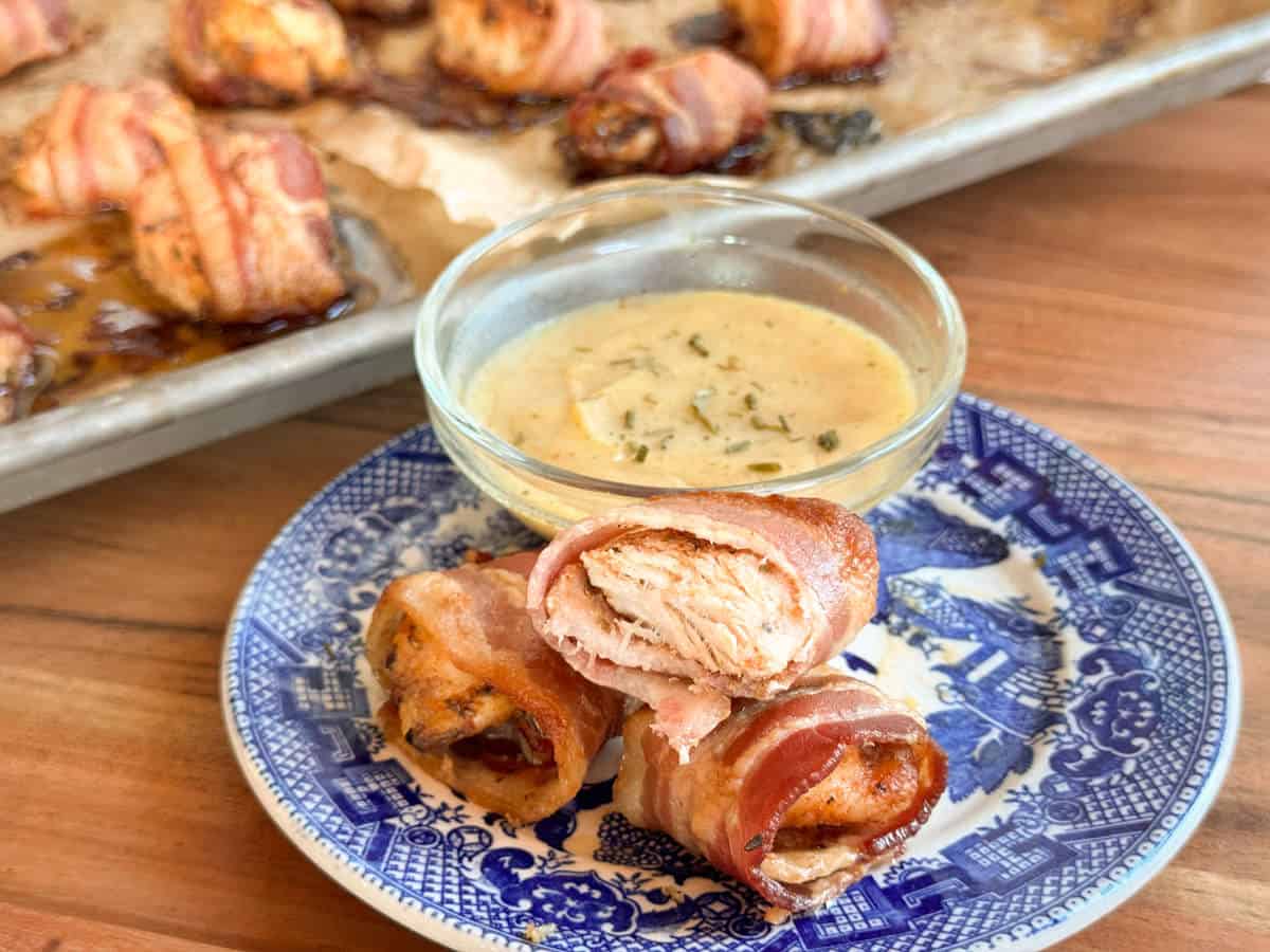Three pieces of bacon-wrapped chicken are on a blue and white plate next to a small bowl of dipping sauce. More bacon-wrapped chicken pieces are on a parchment-lined baking sheet in the background.
