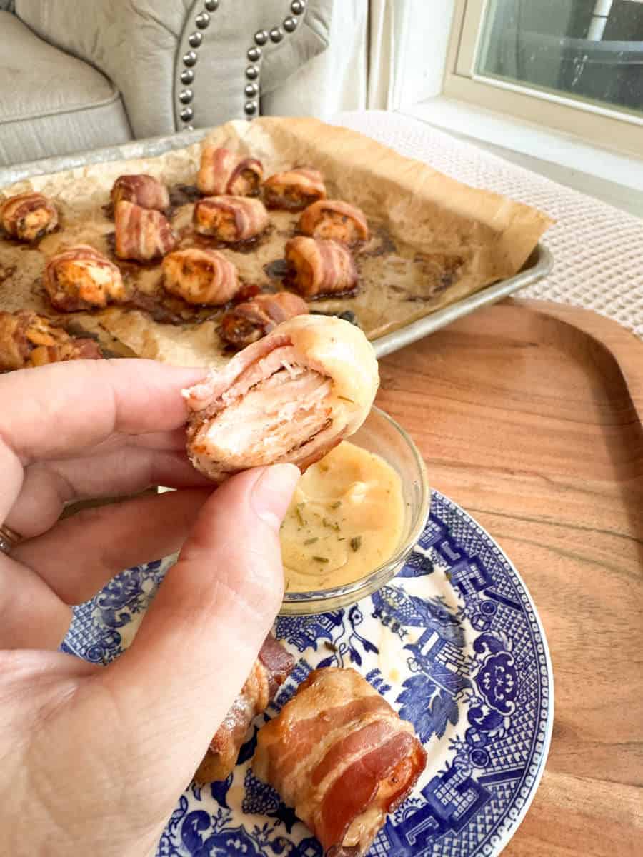 A hand holds a bacon-wrapped chicken bite over a small bowl of dipping sauce on a blue-patterned plate. More bacon-wrapped bites are on a parchment-lined baking sheet in the background.