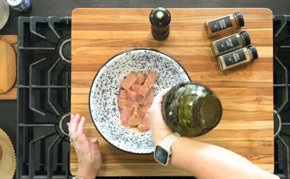 A person pours oil from a bottle into a bowl of raw chicken pieces on a wooden cutting board, with spice jars and a pepper grinder nearby on a stovetop.
