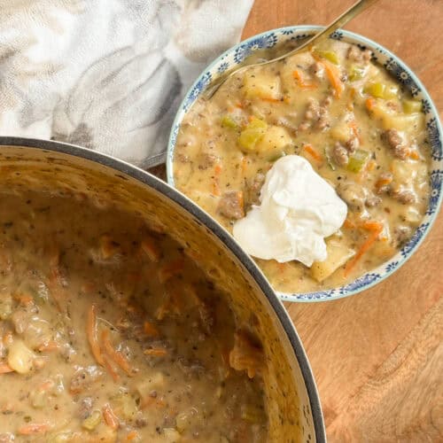 A bowl of thick, creamy soup with visible chunks of potato, carrot, celery, and ground meat, topped with a dollop of sour cream, sits beside a pot containing more of the same soup on a wooden surface.