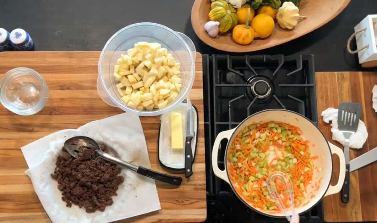 A kitchen scene showing cooked ground meat on a plate, a bowl of diced potatoes, butter, and a pot of chopped carrots and celery cooking on a stove. Decorative squash are on a dish nearby. Kitchen utensils are visible.