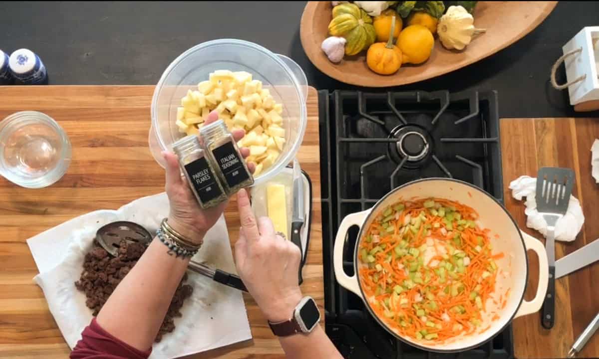 A person holding a bowl of chopped apples and two spice jars over a wooden cutting board with cooked ground meat, near a stovetop with a pot of sautéing carrots and celery, and a bowl of assorted squash.